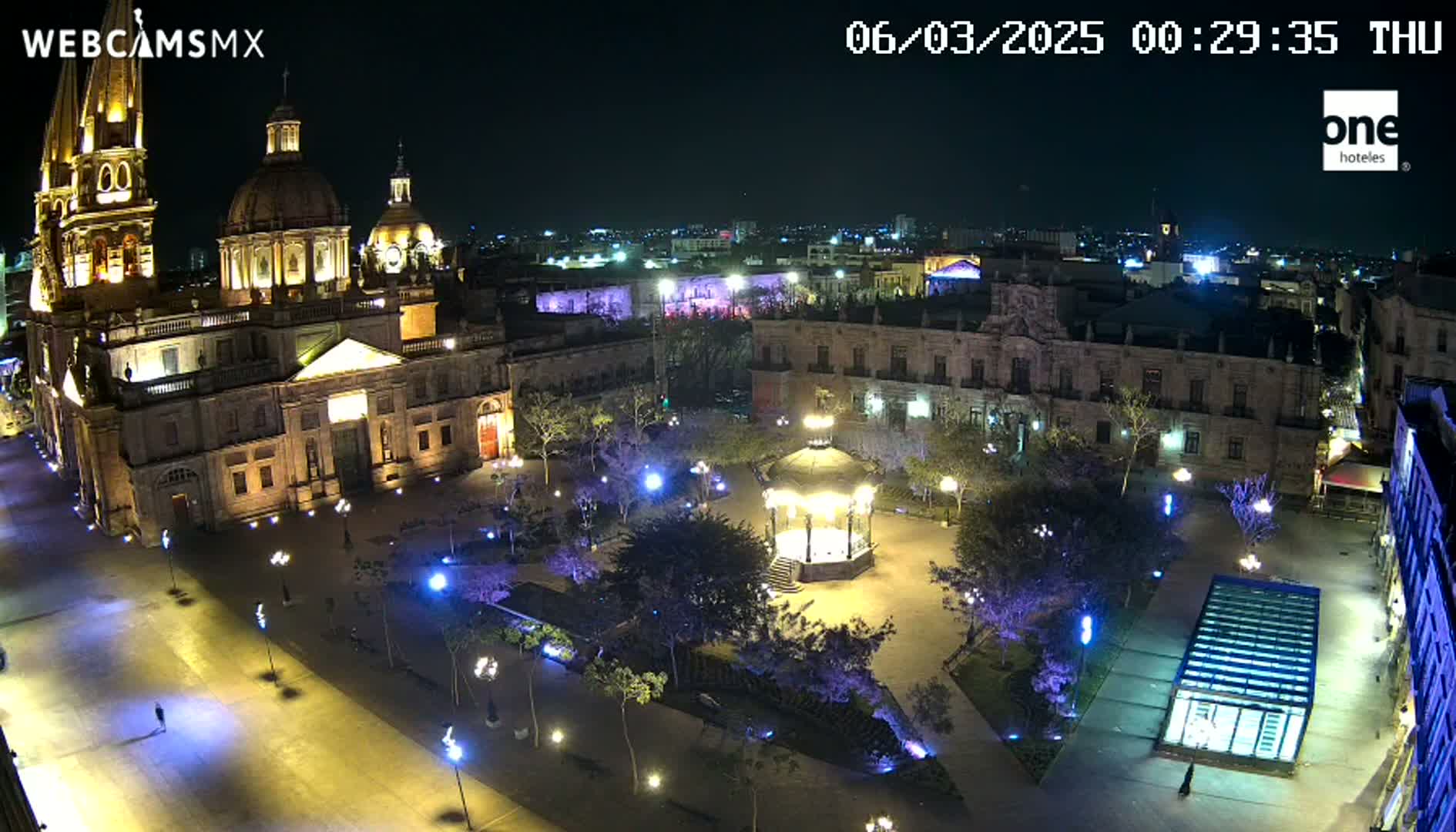 A brightly lit plaza at night, featuring a large cathedral, a gazebo, and surrounding buildings, under a clear, dark sky.