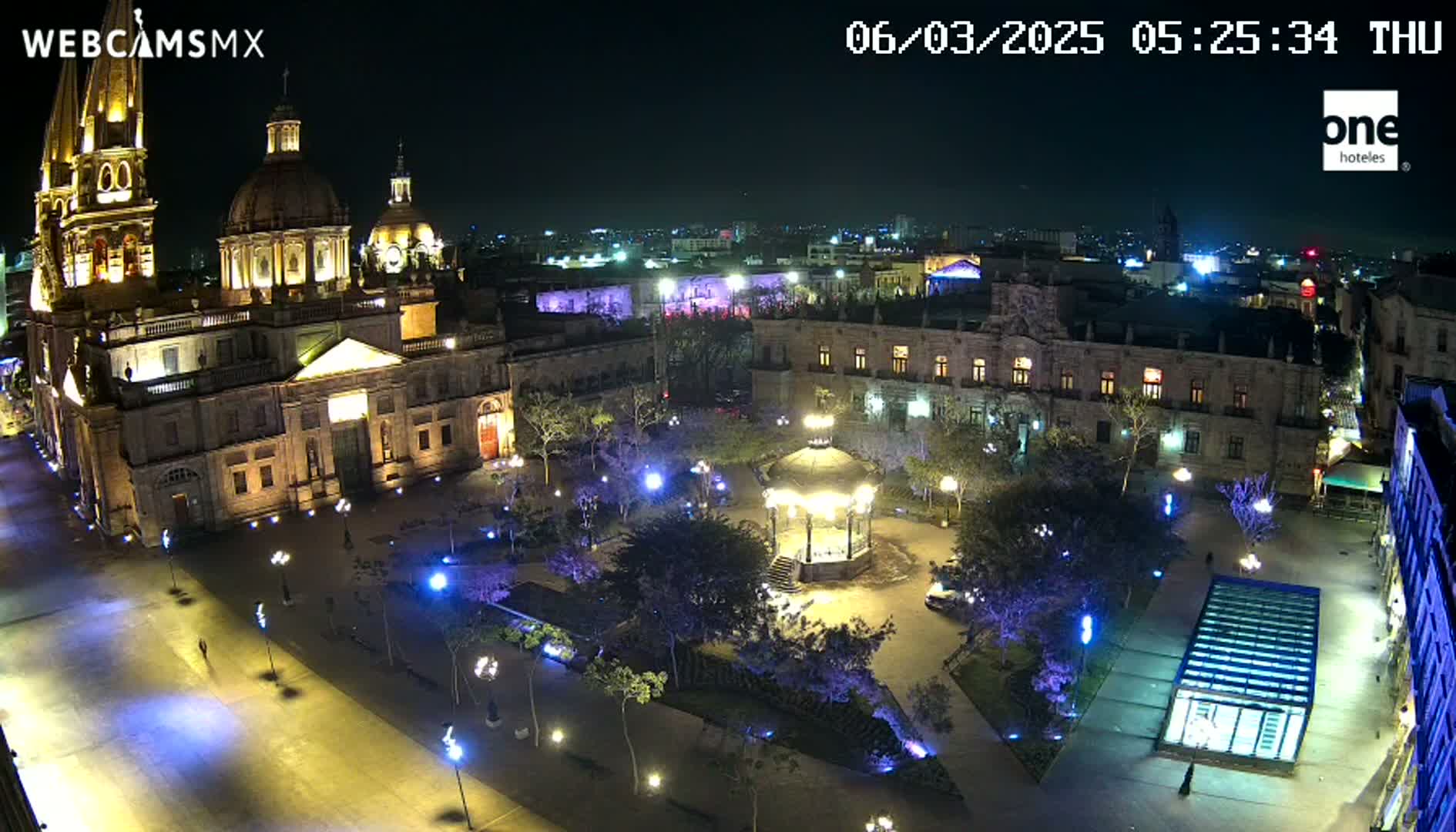 A brightly lit plaza at night, centered around a gazebo, with a large cathedral and other buildings visible in the background under a clear, dark sky.