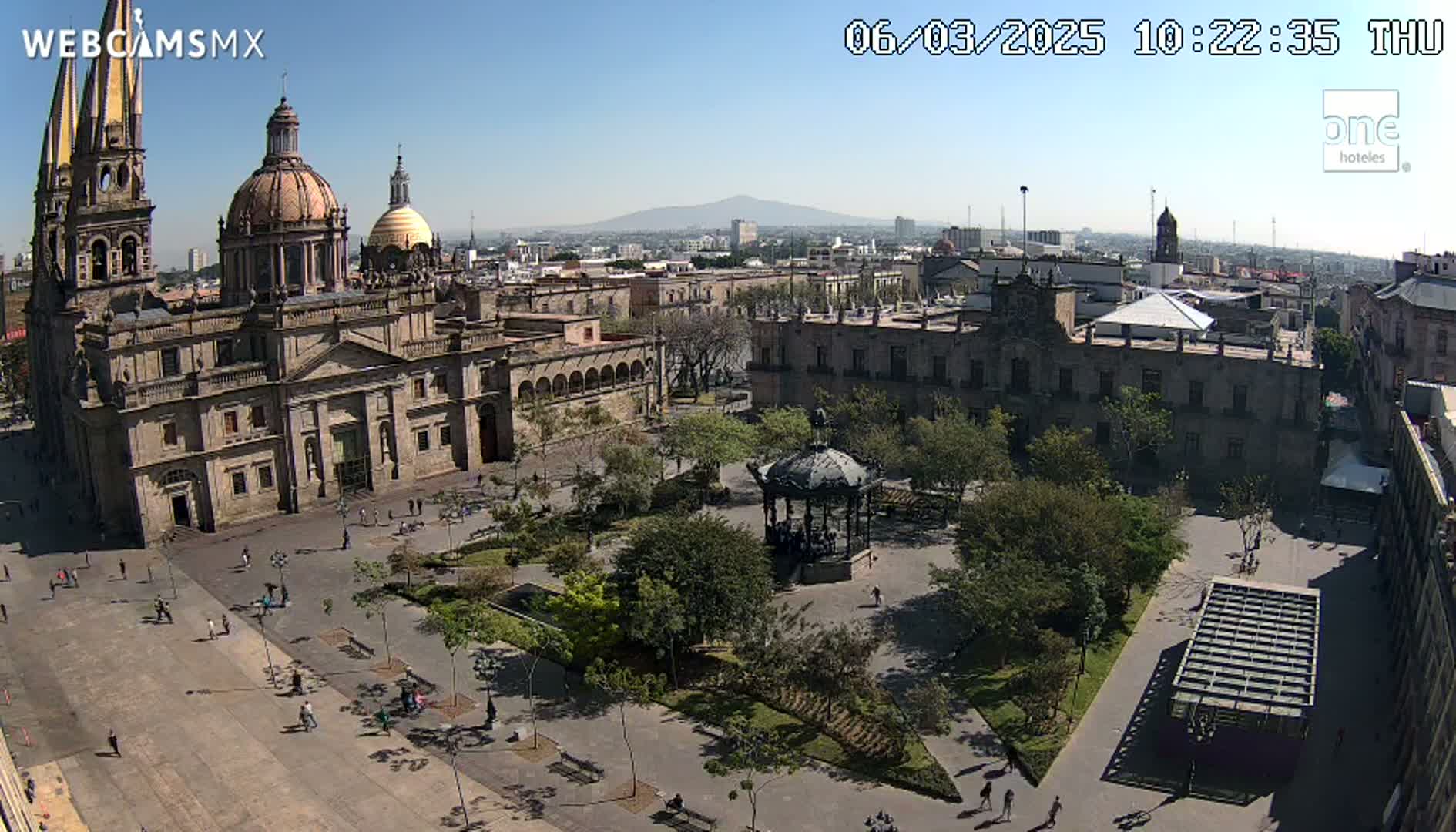 A large plaza with a cathedral, government buildings, a gazebo, and many trees is seen on a sunny day with a distant mountain visible in the background.