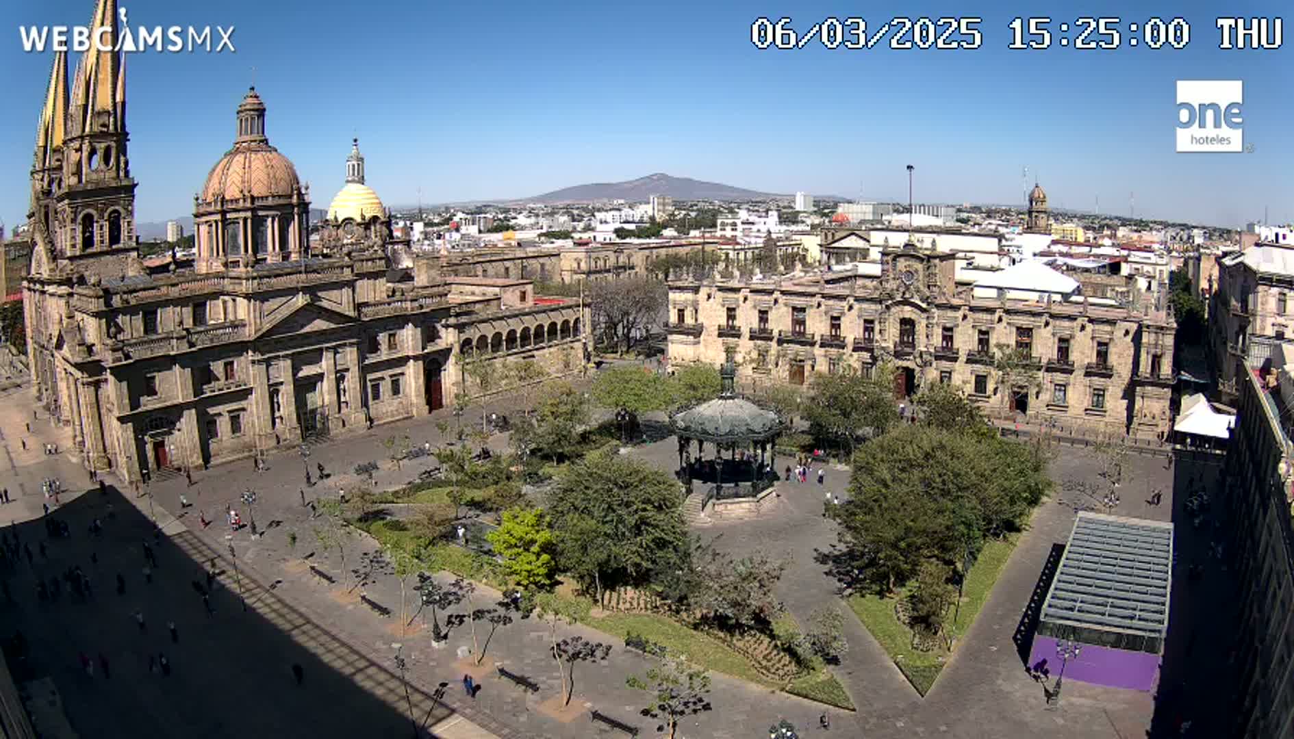 A large plaza with a cathedral and government buildings is shown on a sunny day, with a gazebo in the center of the plaza and many people walking around.