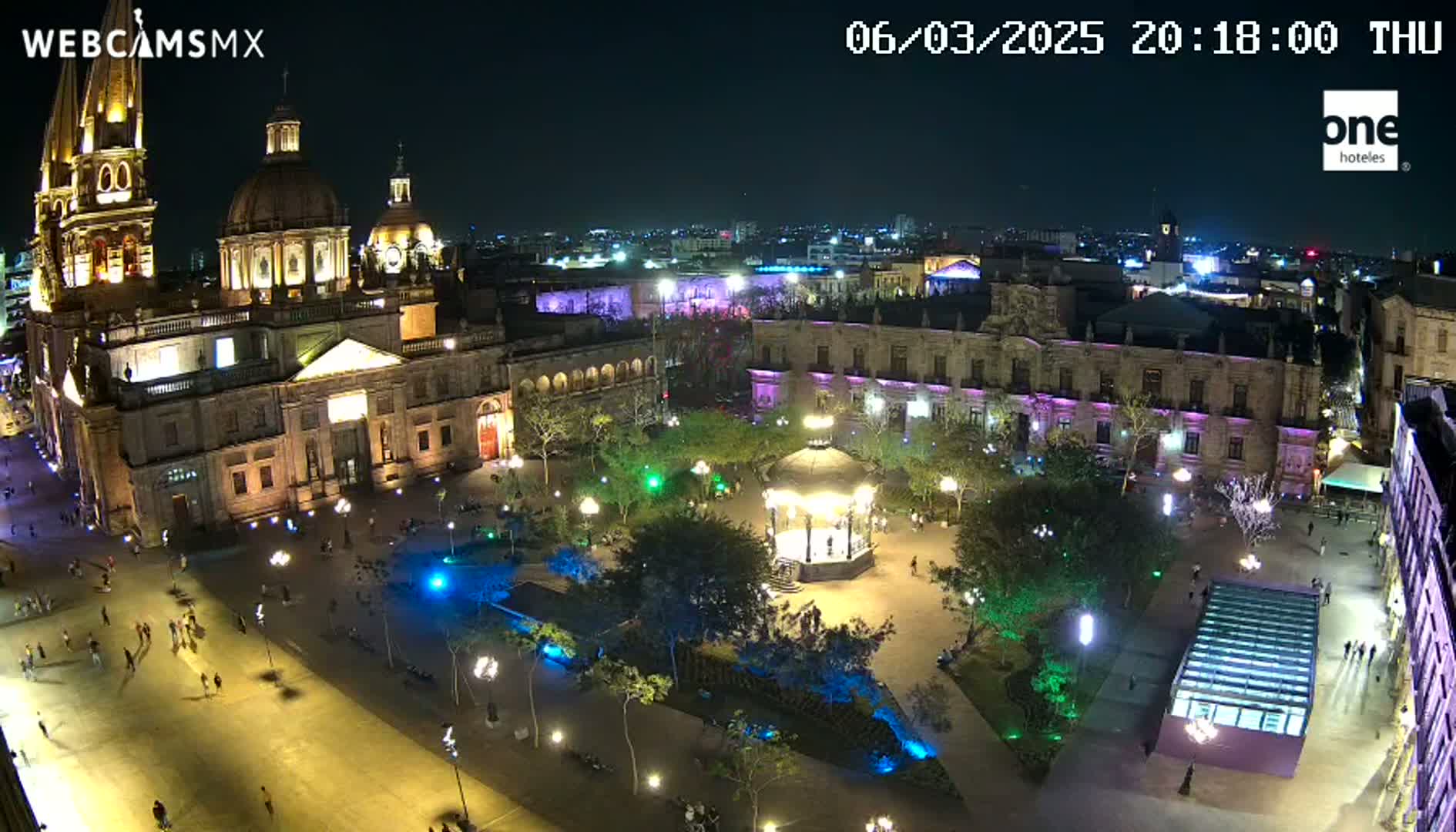 A brightly lit plaza at night, featuring a large cathedral, surrounding buildings, a gazebo, and people walking around.