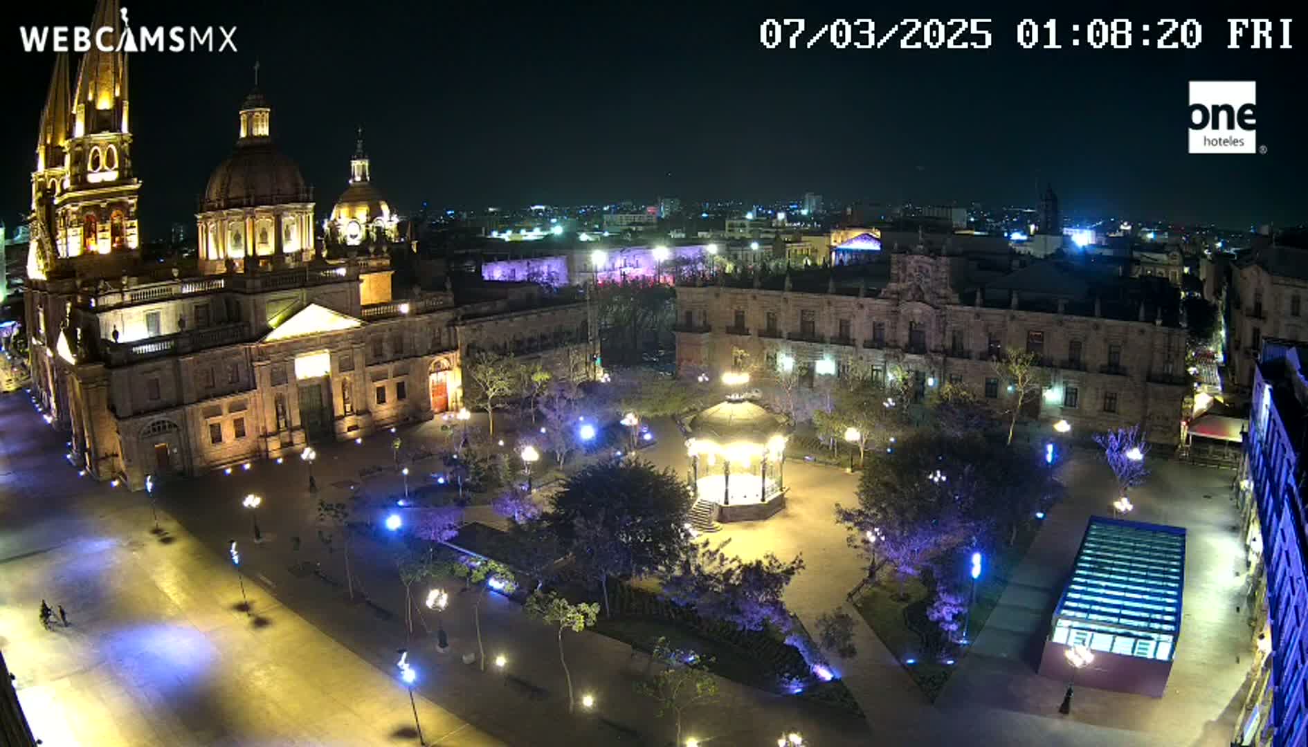 A brightly lit plaza at night, featuring a large cathedral and a gazebo, is surrounded by buildings under a clear, dark sky.