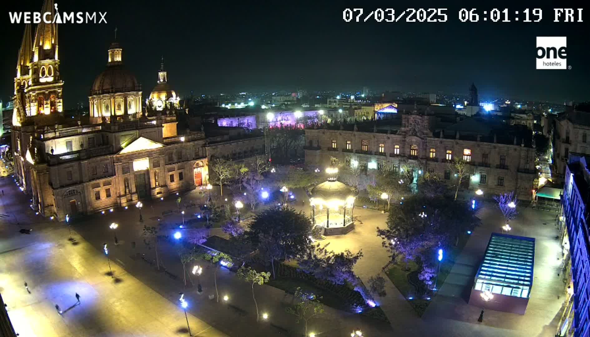 A brightly lit plaza at night features a large cathedral, surrounding buildings, a gazebo, and numerous trees.