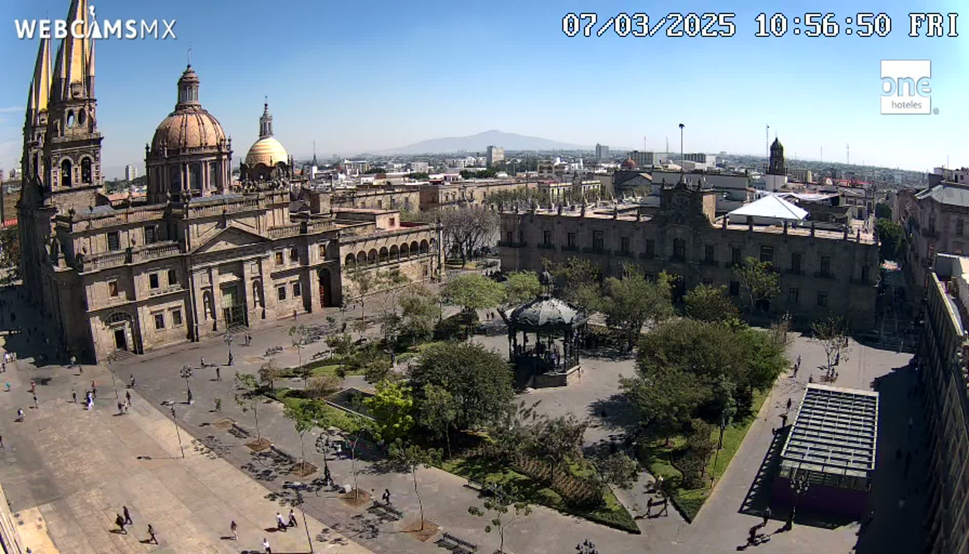 A large plaza with a cathedral, government buildings, a gazebo, and many trees under a clear, sunny sky.