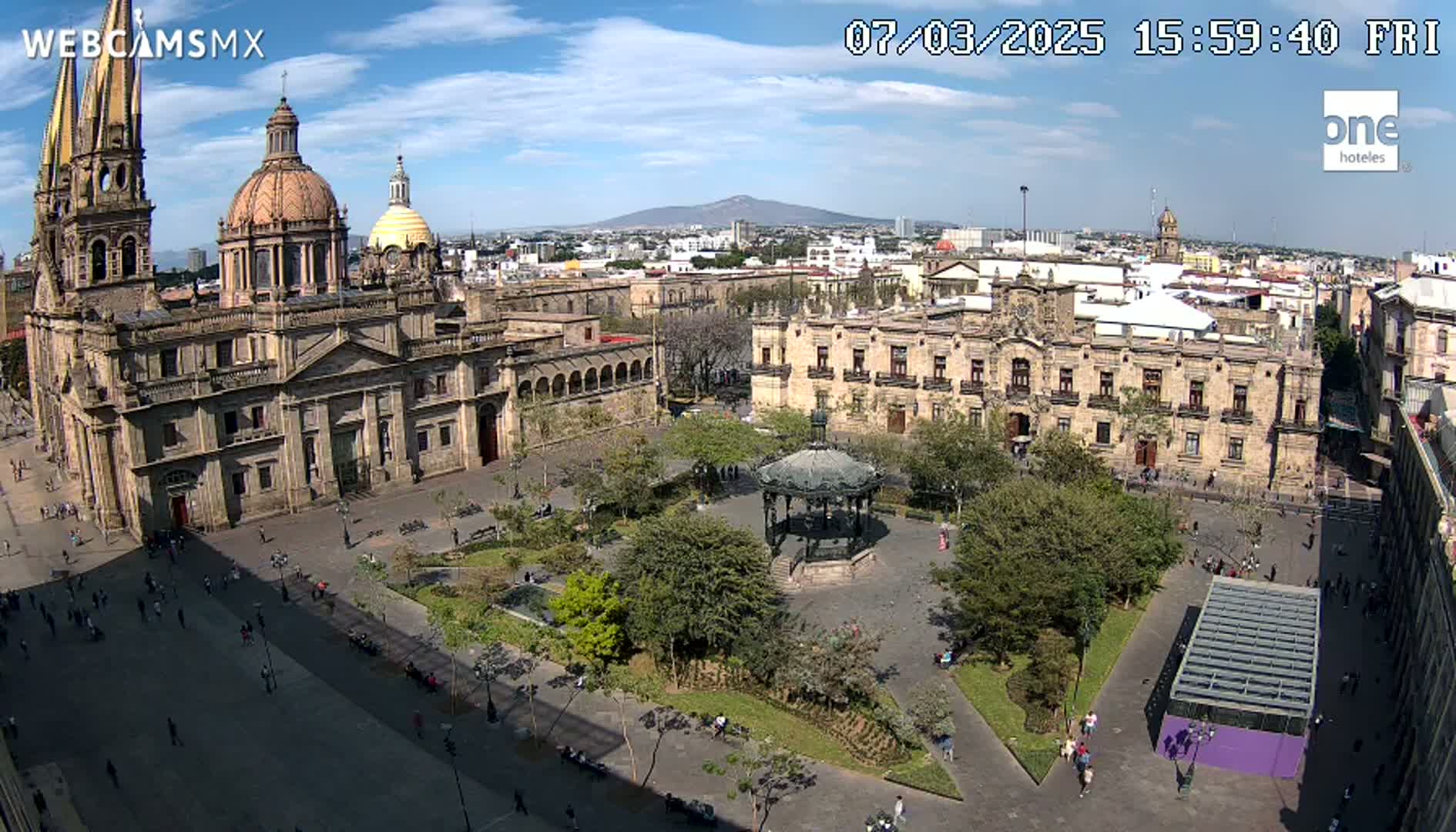 A large plaza with a cathedral, government buildings, a gazebo, and trees is seen under a mostly sunny sky.