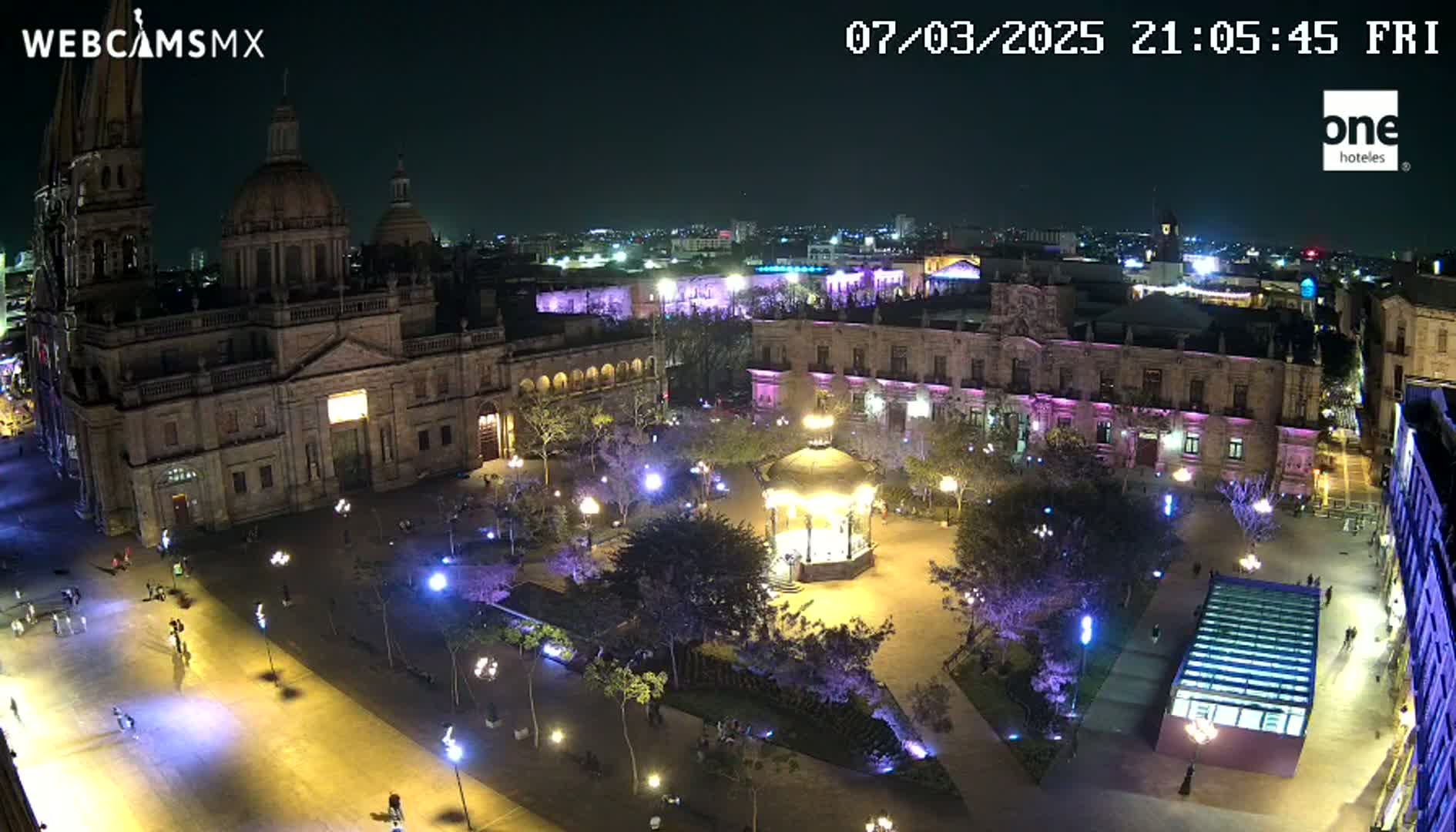 A brightly lit plaza at night, featuring a large cathedral and surrounding buildings, with a gazebo at its center.