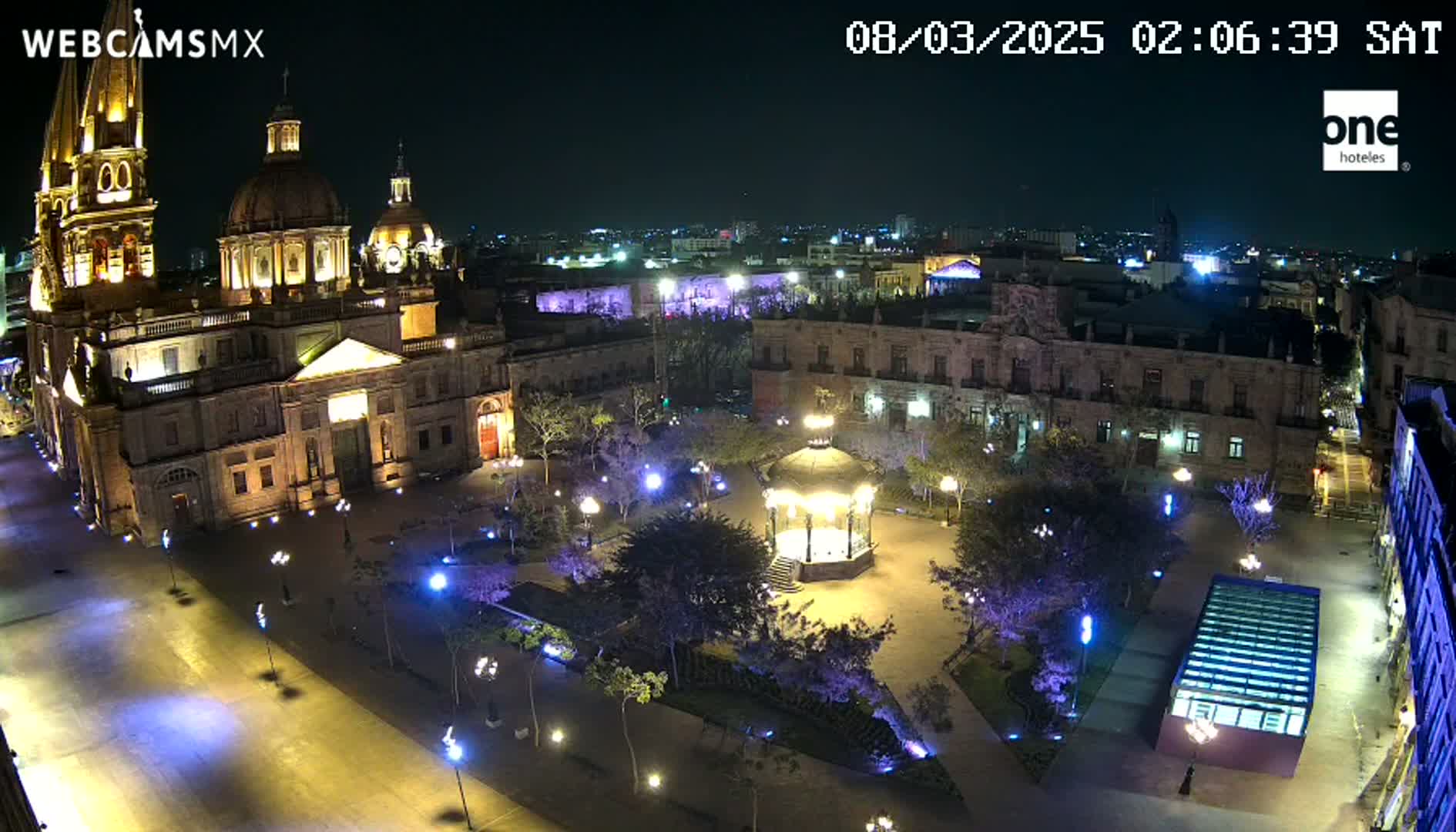 A brightly lit plaza at night, featuring a large cathedral, a gazebo, and surrounding buildings, under a clear, dark sky.
