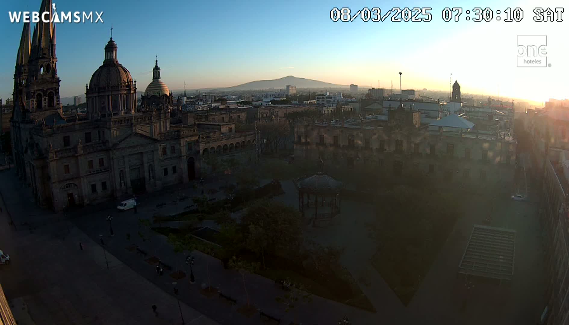 A large cathedral sits in a city square at sunrise, with a distant volcano visible on a clear, bright morning.