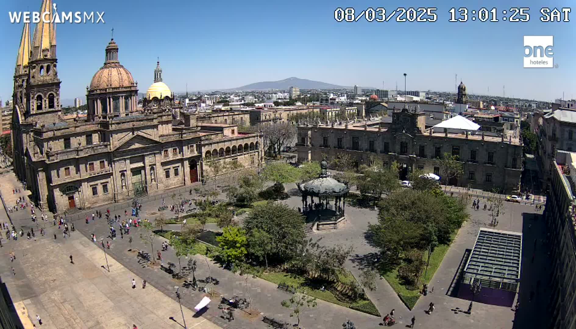 A large plaza with a gazebo, surrounded by buildings including a cathedral, under a clear sunny sky.