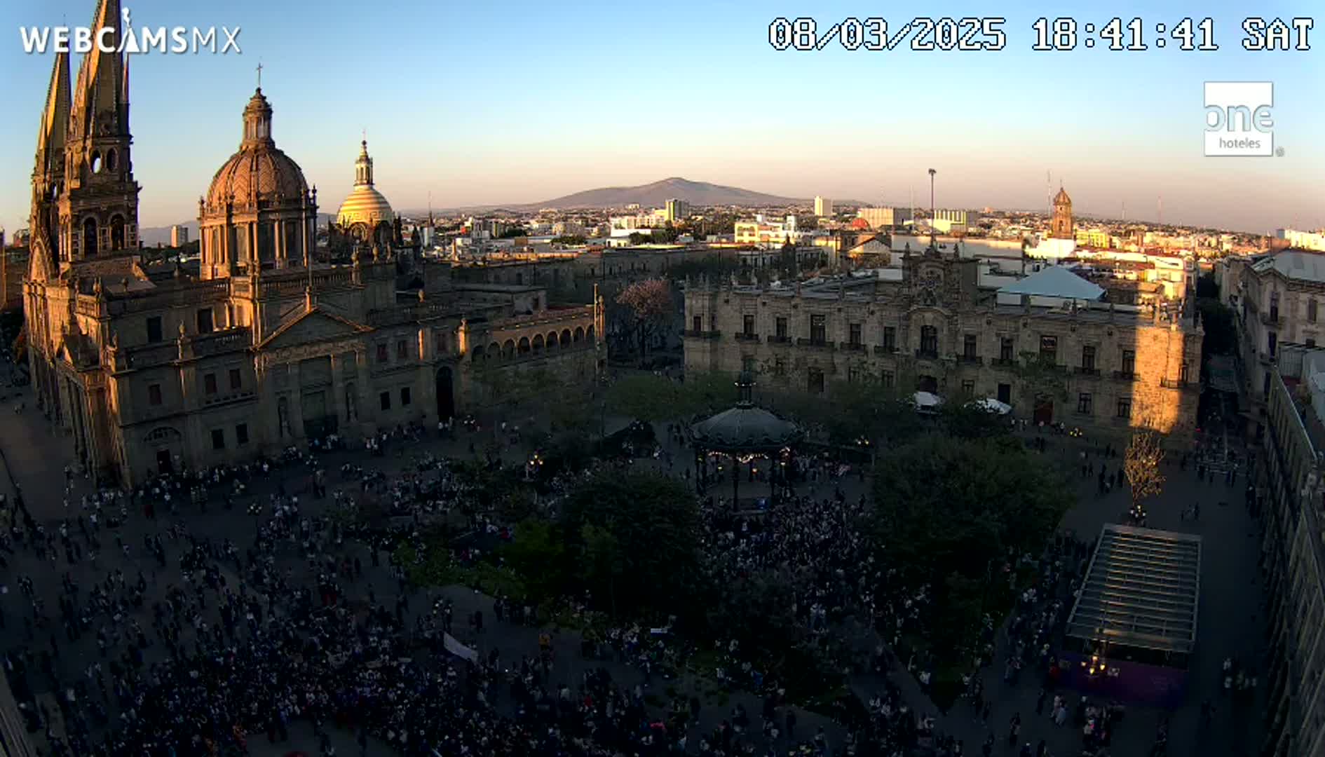 A large crowd gathers in a city square at sunset, with a cathedral and other buildings visible in the background under a clear sky.