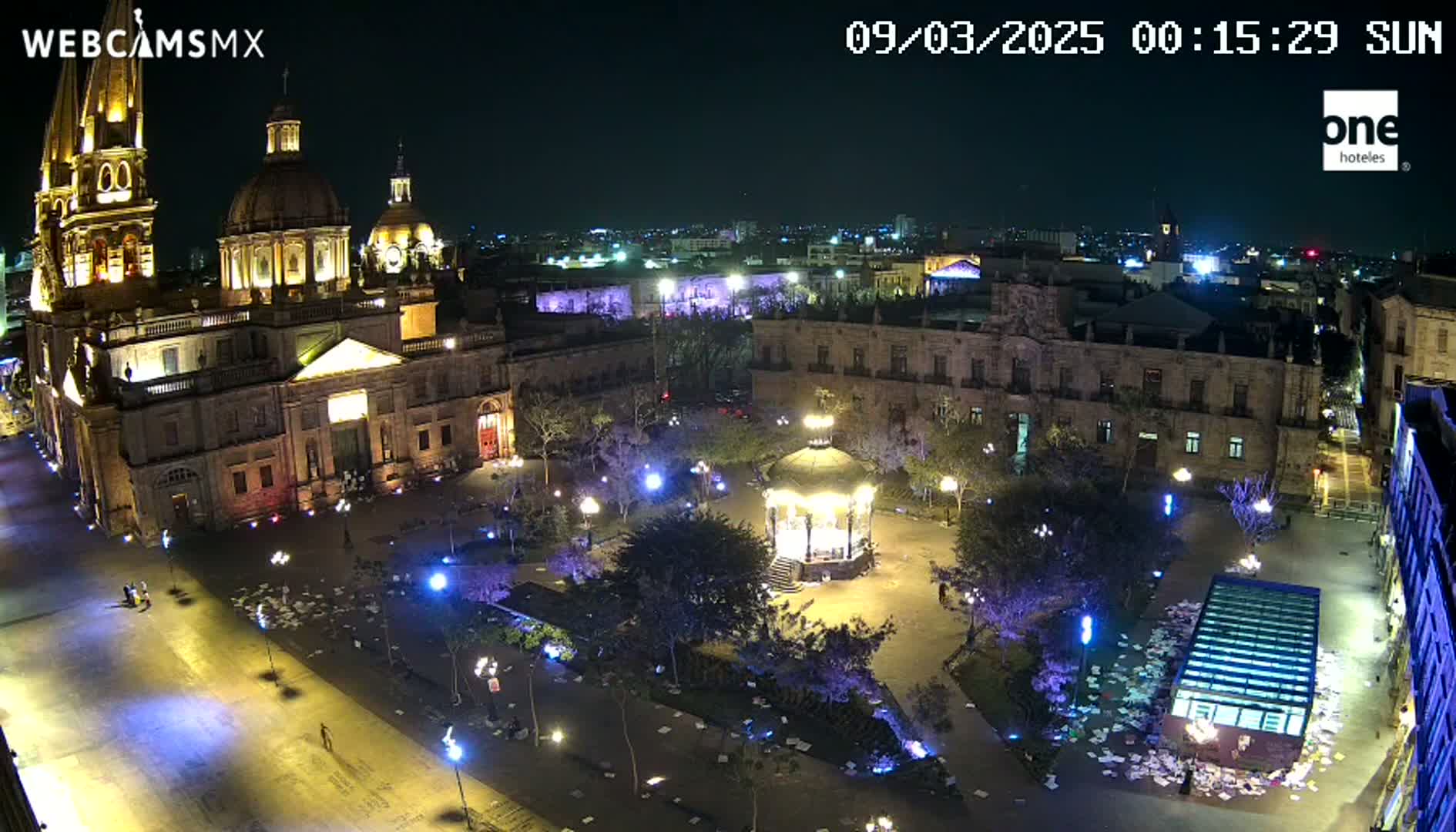 A brightly lit plaza at night, featuring a large cathedral and a gazebo, is strewn with scattered papers under a clear sky.