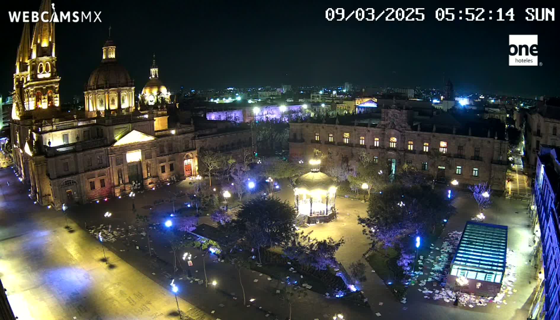 A nighttime, aerial view of a plaza with a large cathedral and surrounding buildings, illuminated by lights, and scattered debris on the ground.