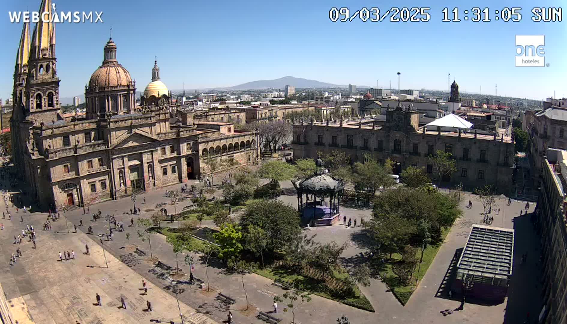 A large plaza with a cathedral and other buildings, a gazebo, and many trees, on a sunny day with a mountain visible in the distance.