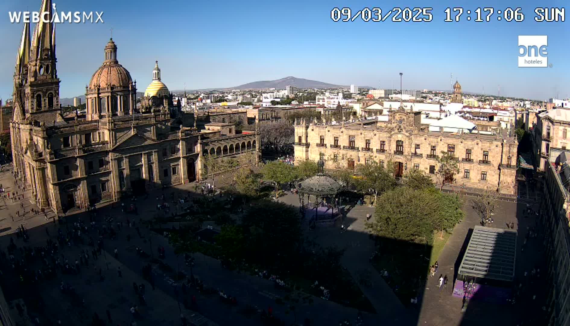 A high-angle, sunny view shows a large plaza with a cathedral and government building, surrounded by other buildings and trees under a clear sky.
