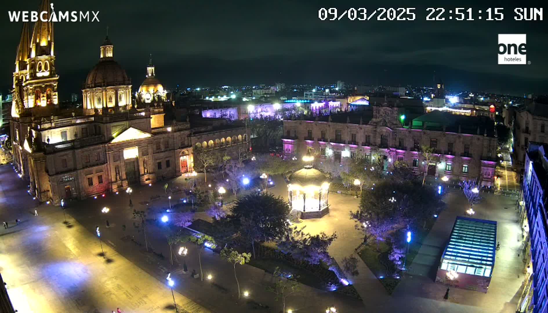 A brightly lit plaza at night, centered around a gazebo, with a large cathedral and other buildings visible in the background under a clear, dark sky.