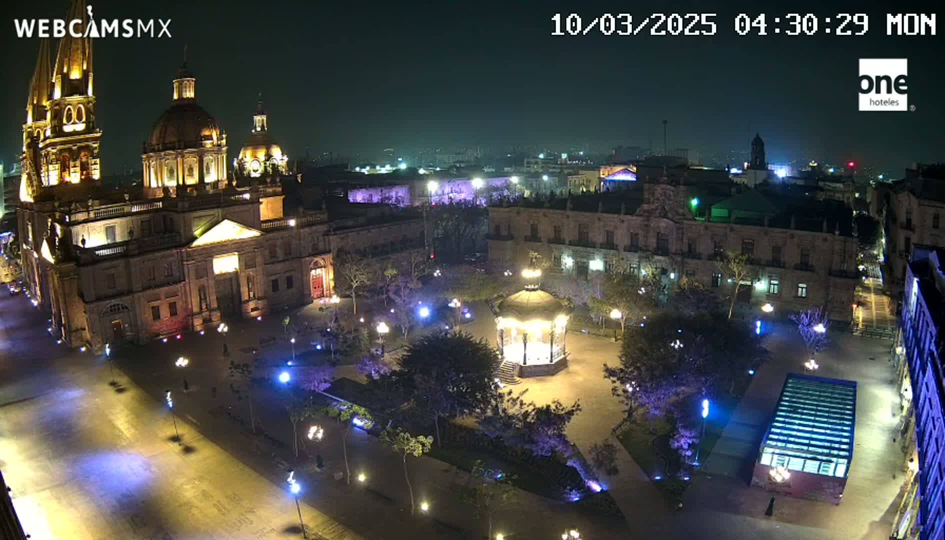 A brightly lit plaza at night features a large cathedral, surrounding buildings, a gazebo, and numerous trees and lights.