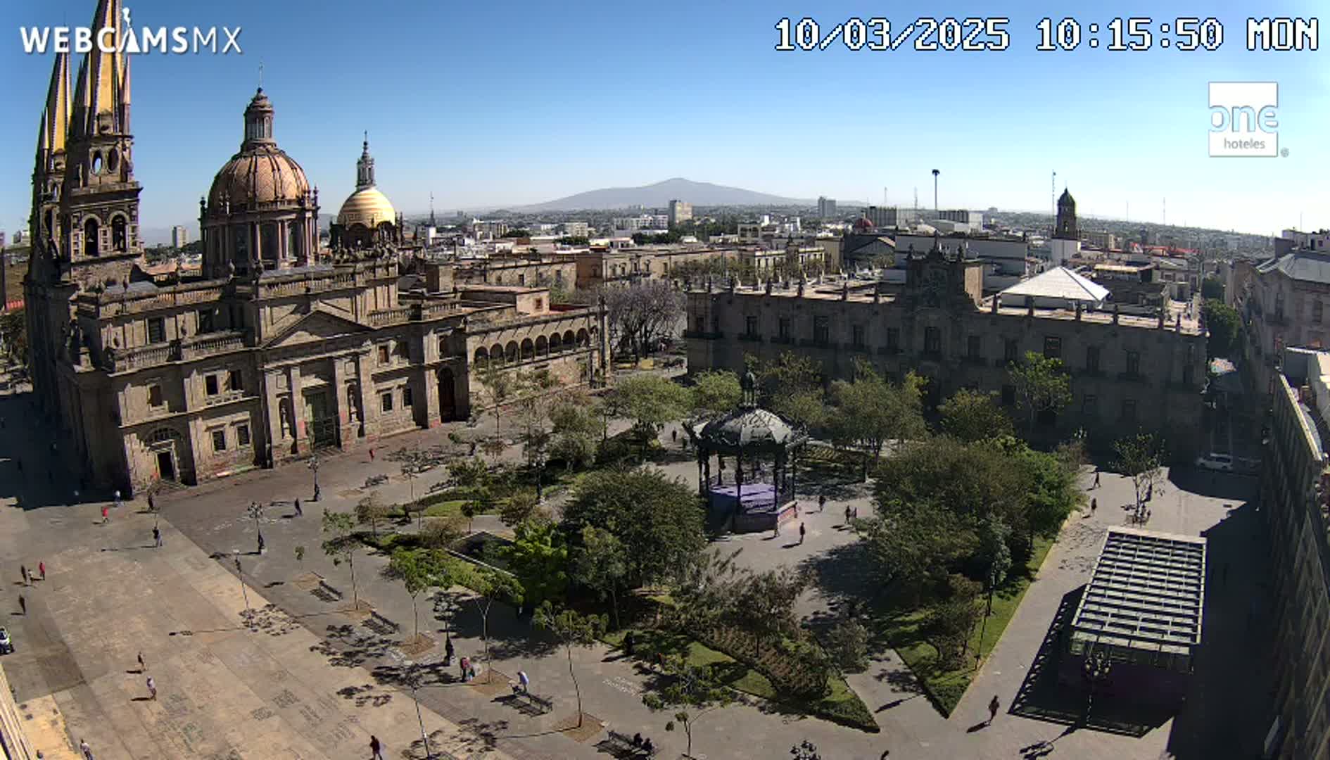 A large plaza with a cathedral, government buildings, a gazebo, and many trees is visible under a clear, sunny sky, with a distant mountain in the background.