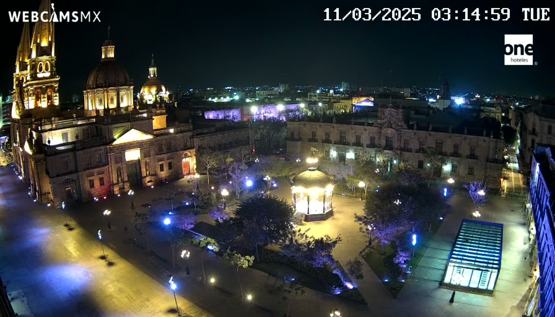 A brightly lit plaza at night, centered around a gazebo, with a large cathedral and surrounding buildings visible under a clear, dark sky.