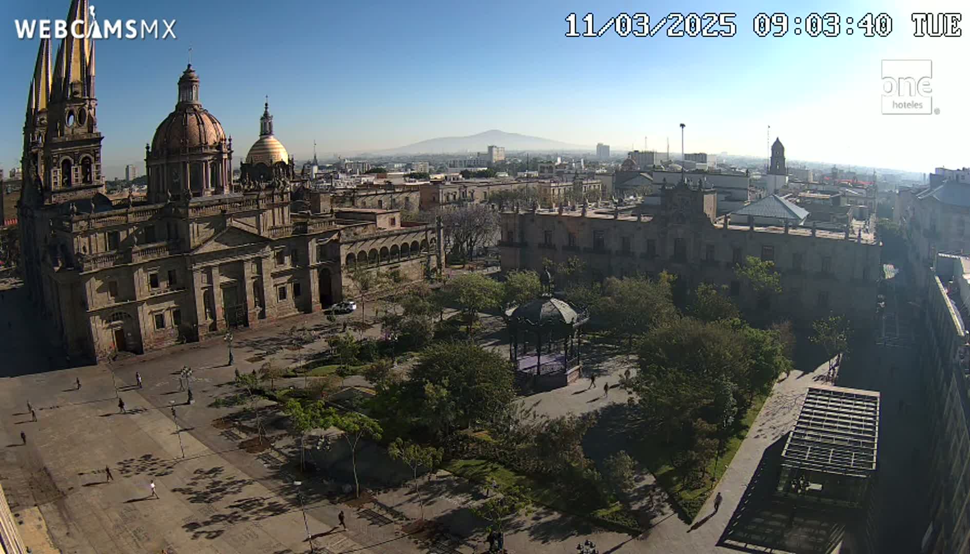A large cathedral and plaza with a gazebo are seen on a sunny day, with a city and distant mountains in the background.