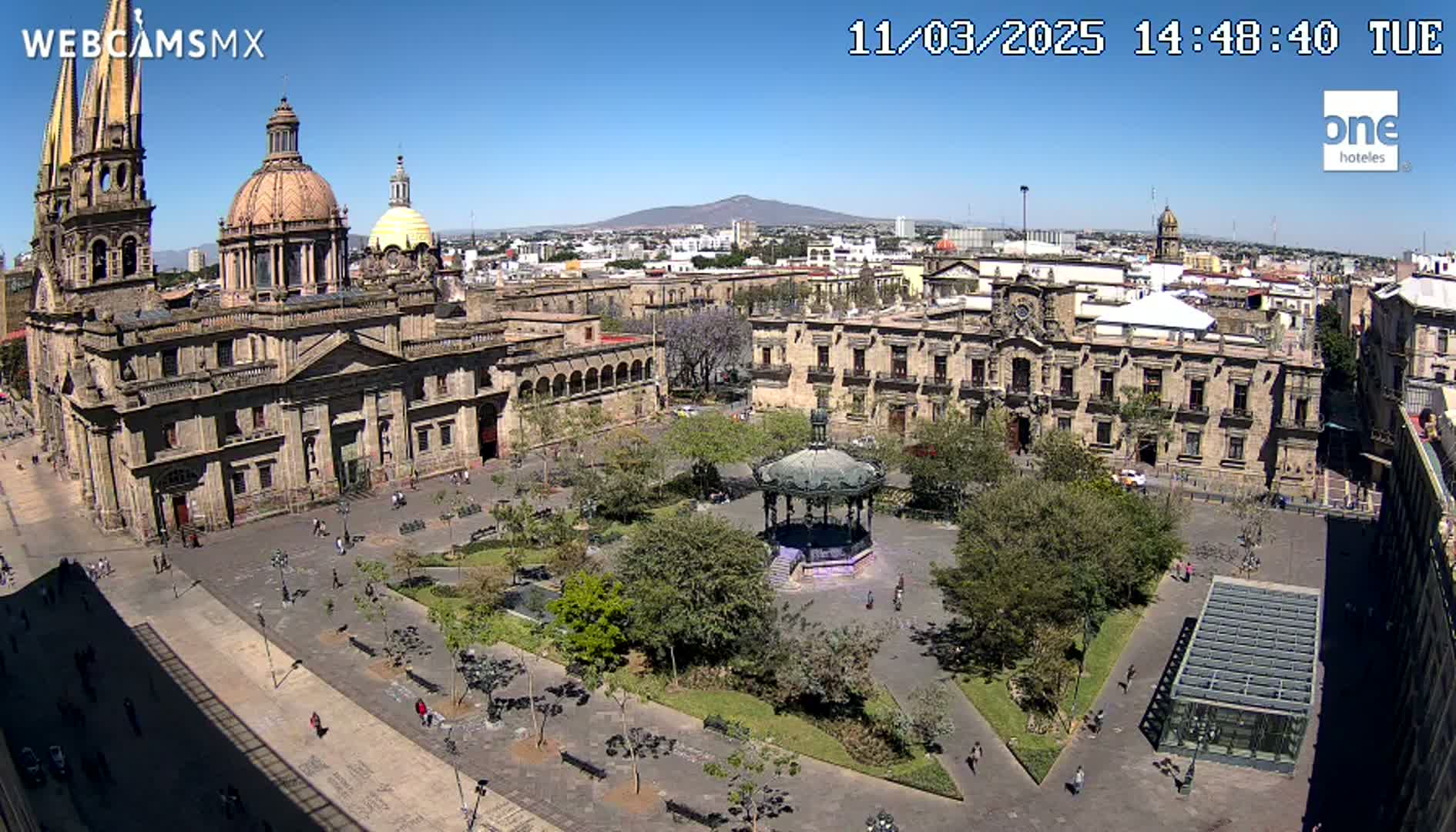 A sunlit city square with a large cathedral, government buildings, a gazebo, and trees under a clear blue sky.