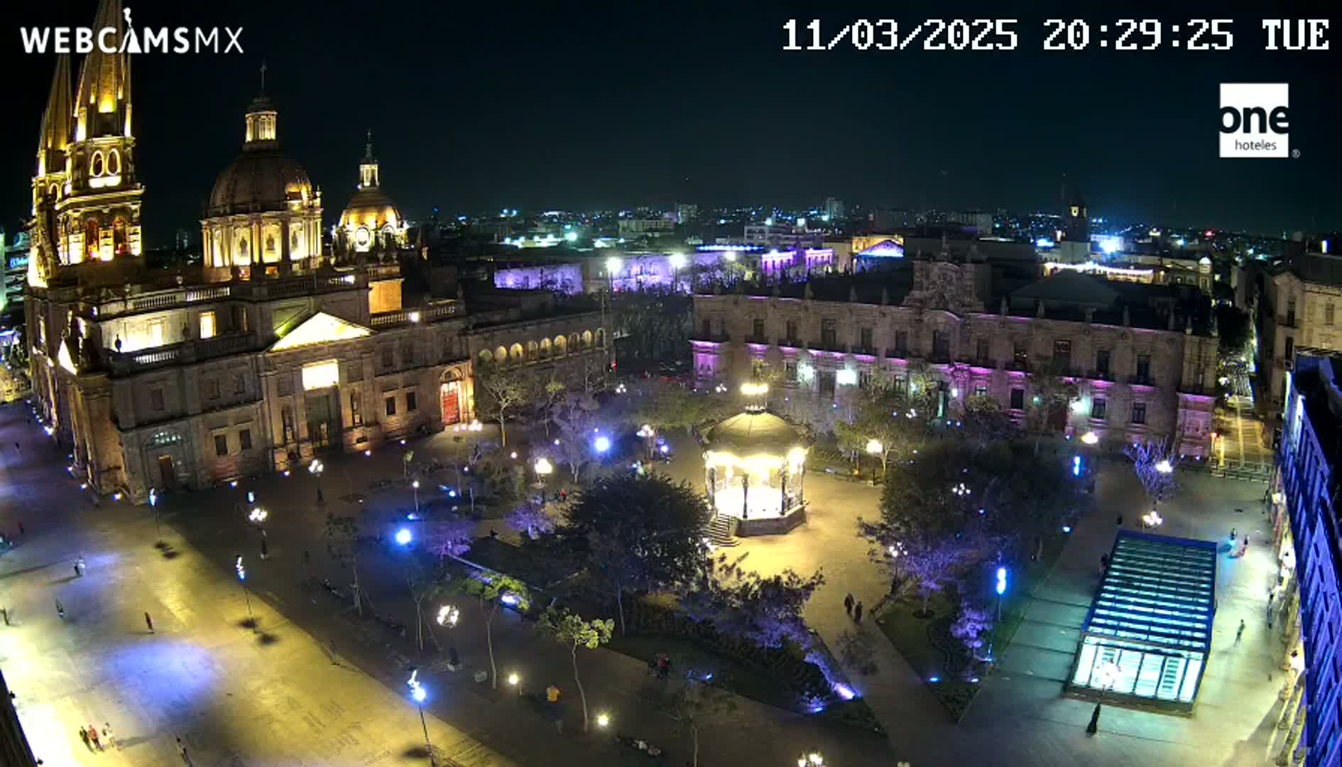 A brightly lit plaza at night, featuring a large cathedral and gazebo, surrounded by buildings under a clear, dark sky.