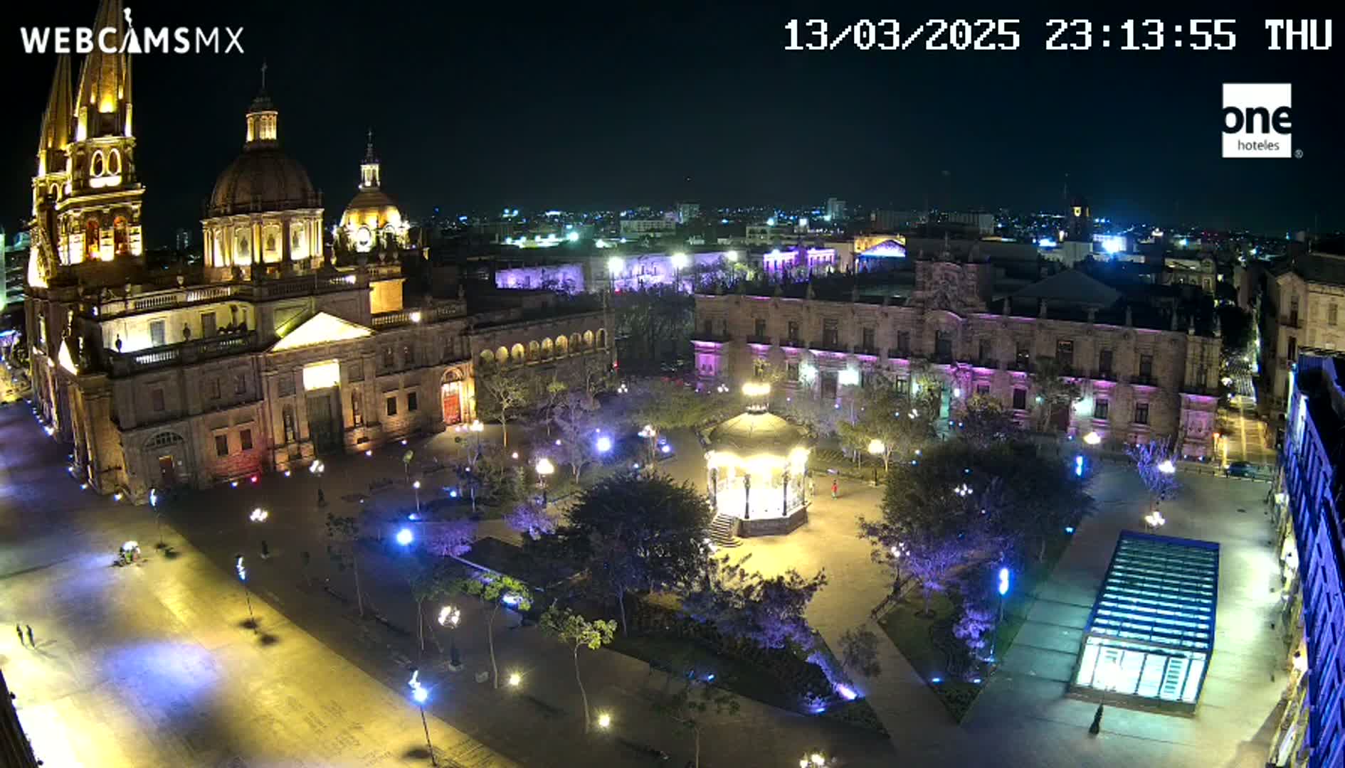 A brightly lit plaza at night, featuring a large cathedral, surrounding buildings, a gazebo, and trees, under a clear, dark sky.