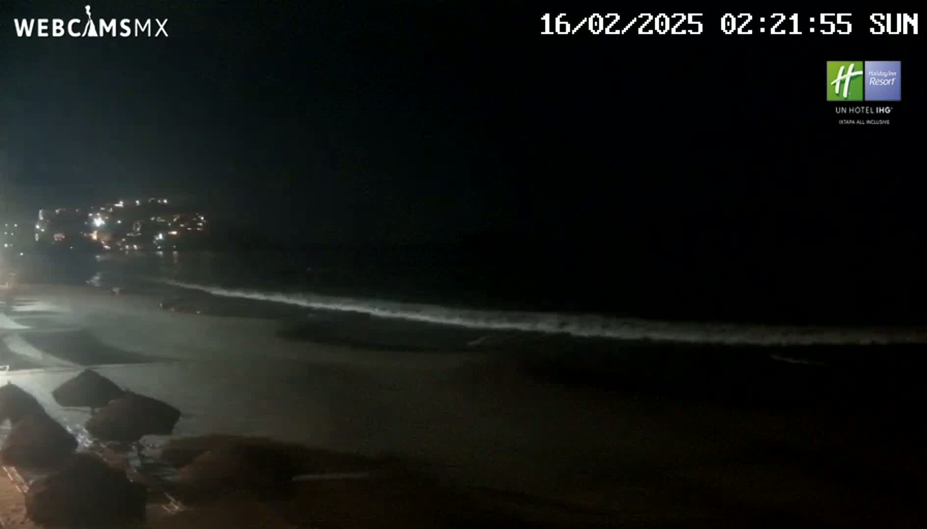 A dark nighttime beach scene shows gentle waves lapping the shore under a clear sky, with lights from buildings visible in the distance.