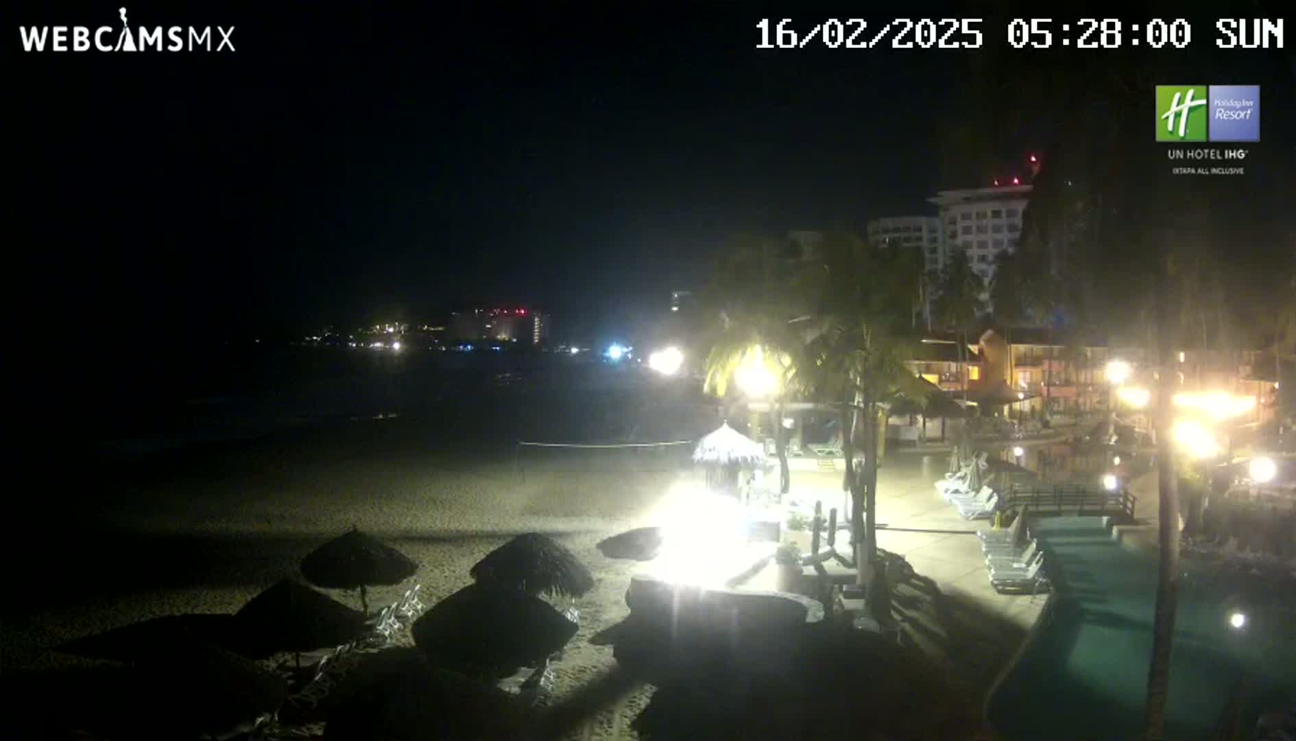A nighttime beach scene shows a sandy shore with beach umbrellas, palm trees, resort buildings, and a swimming pool under a clear, dark sky.