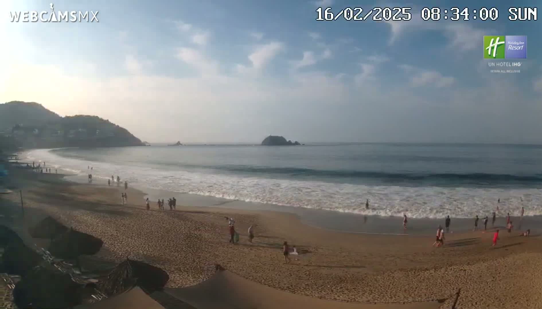 A sandy beach with scattered people is visible, alongside a calm ocean under a partly cloudy sky.