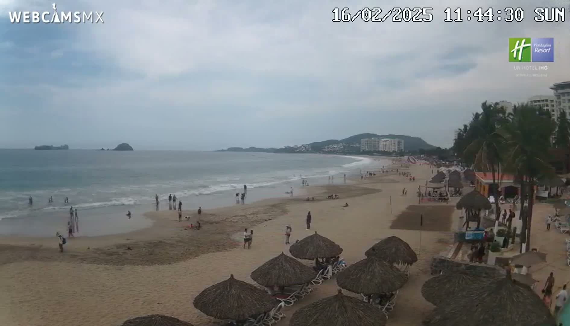 A sandy beach with a calm ocean, some people, and thatch-roofed beach umbrellas under an overcast sky.