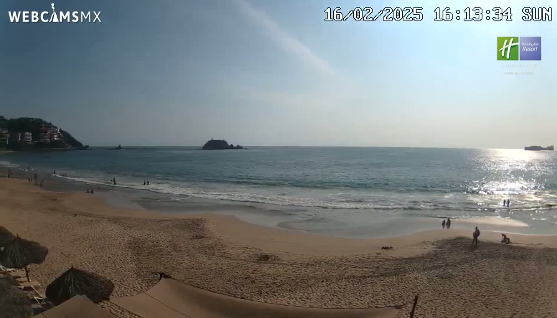 A sandy beach with a few people on it slopes gently down to a calm ocean under a mostly clear, sunny sky.