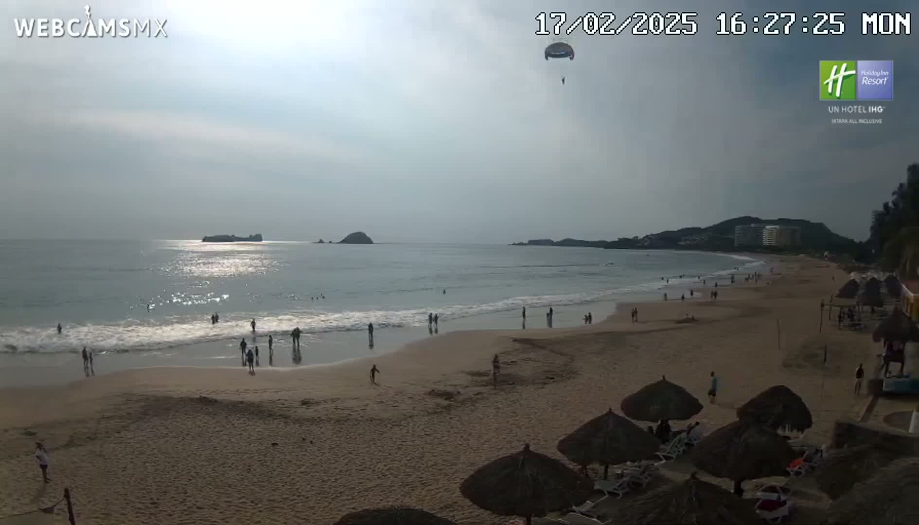 A sandy beach with people swimming and sunbathing under a mostly cloudy sky, with a parasail visible in the distance.