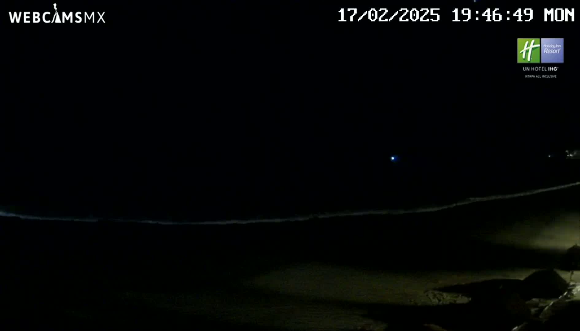 A dark, nighttime beach scene shows a faint line of waves and a distant light in the sky.
