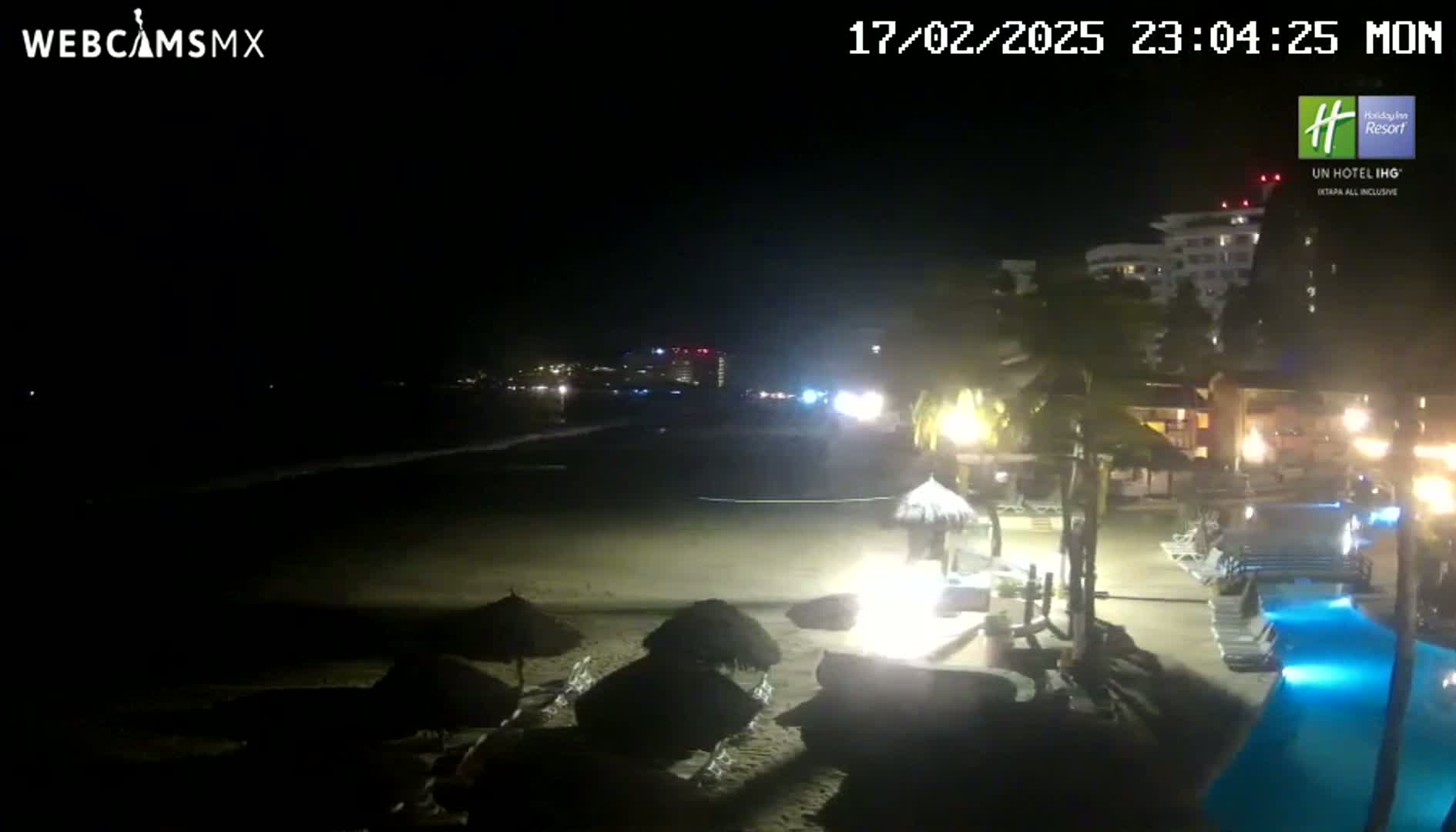 A nighttime beach scene shows a resort with a lit pool and beach umbrellas on a sandy beach, under a clear, dark sky.