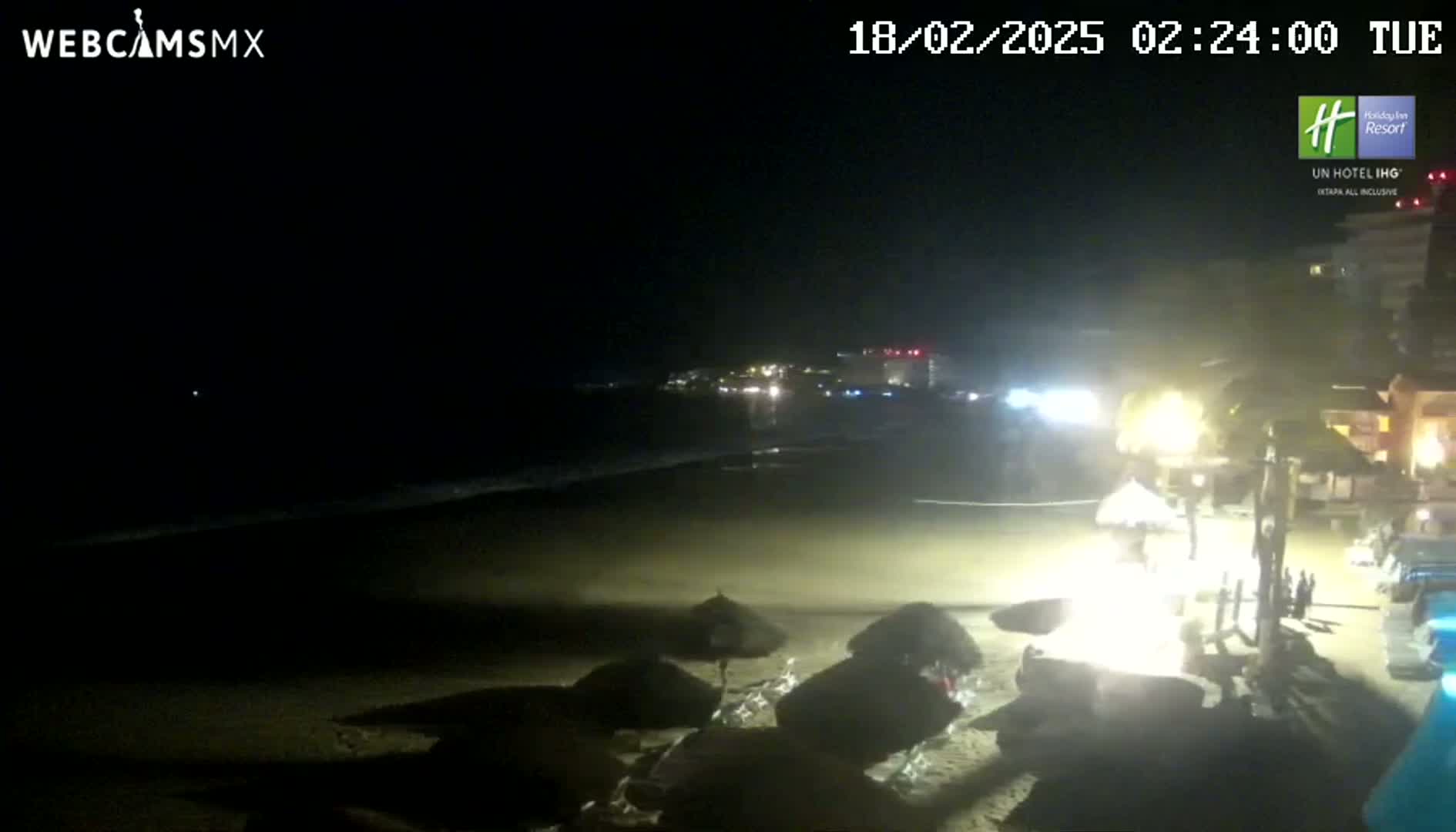 A nighttime beach scene shows dark sand, gentle waves, closed umbrellas, and a dimly lit beachfront building.