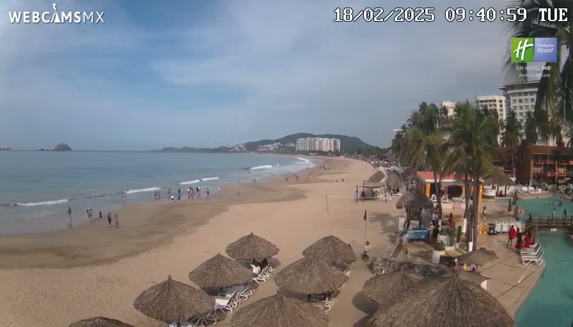 A sandy beach with calm waves, scattered beachgoers, thatch-roofed umbrellas, and palm trees lines a coastline next to a resort on a clear, sunny day.