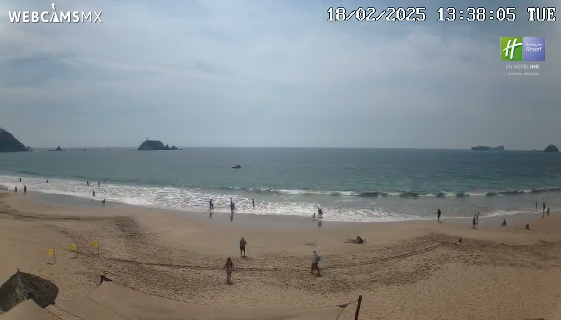 A mostly sunny day at a sandy beach shows calm waves lapping the shore, with a few people swimming and relaxing on the sand.