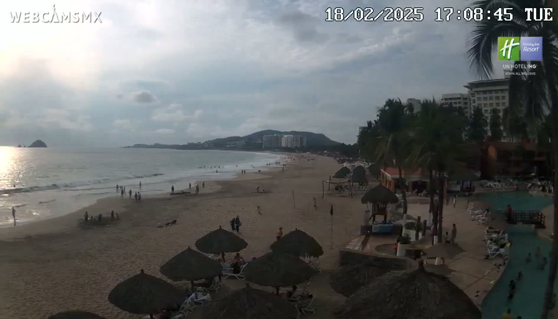 A mostly sunny beach scene shows a sandy shore with scattered beachgoers, palm trees, beach umbrellas, and a hotel in the background, with calm ocean waves lapping the sand.