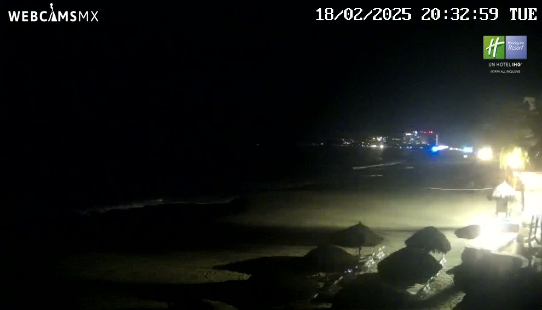 A dark, nighttime beach scene shows several closed umbrellas on the sand near a building, with dimly lit city lights in the distance.
