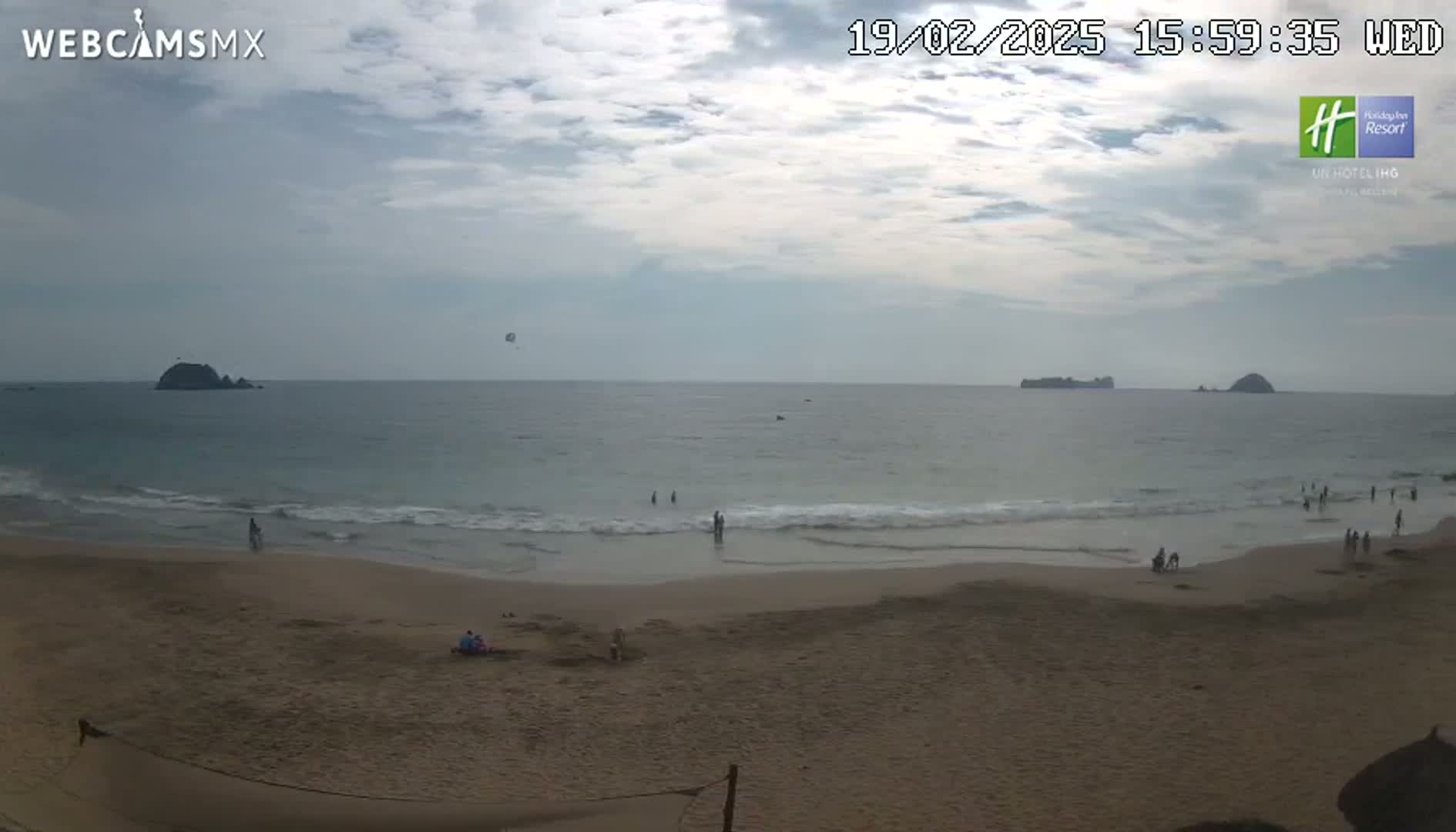 A mostly sunny beach scene shows a calm ocean with small waves lapping a sandy shore where several people are present, with distant rocky islands and a parasail visible in the sky.
