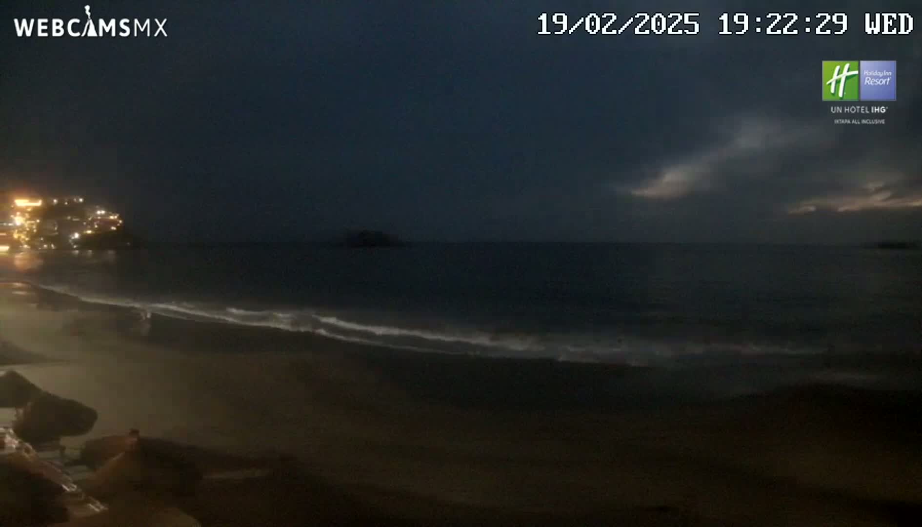 A dark, nighttime beach scene shows a dark ocean with gentle waves lapping onto the shore under a mostly cloudy sky.