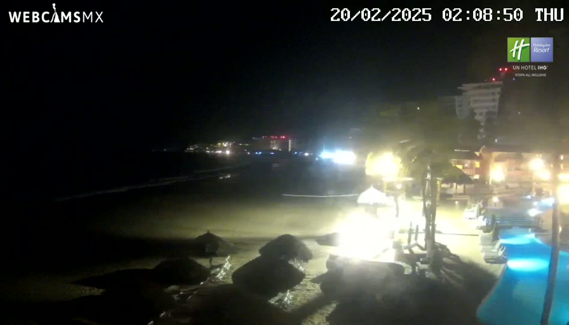A nighttime beach scene shows dark ocean waves lapping a sandy shore, with dimly lit beach umbrellas and resort buildings in the background.