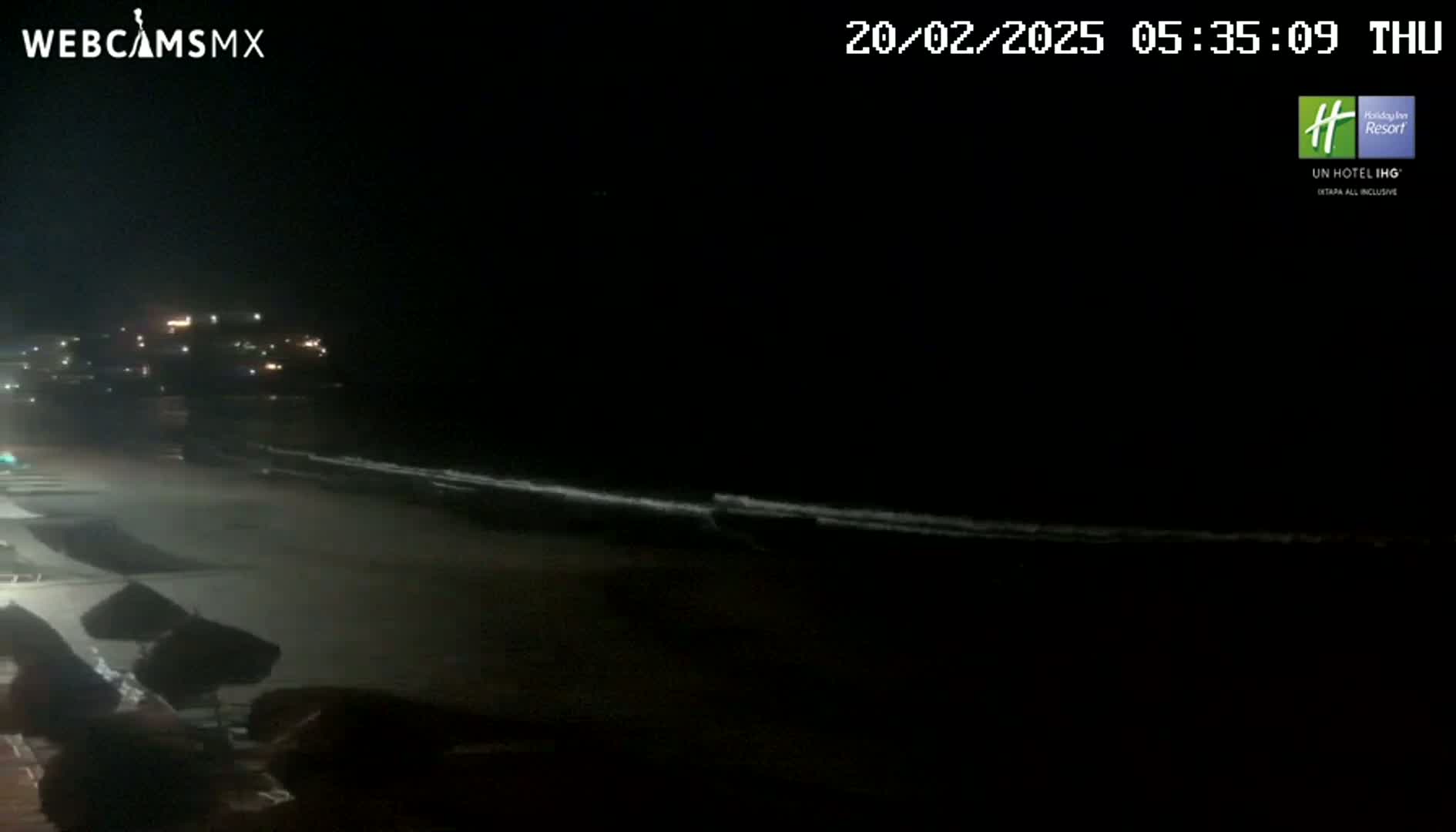 A nighttime beach scene shows dark sand, gently lapping waves, and lights from buildings in the distance.