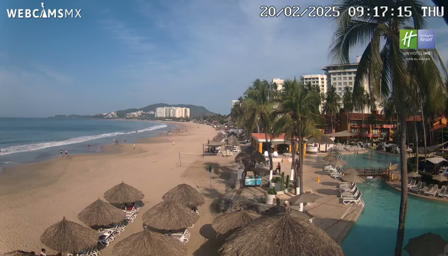 A sandy beach with calm ocean waves, palm trees, resort buildings, and numerous thatched umbrellas is visible on a sunny day.