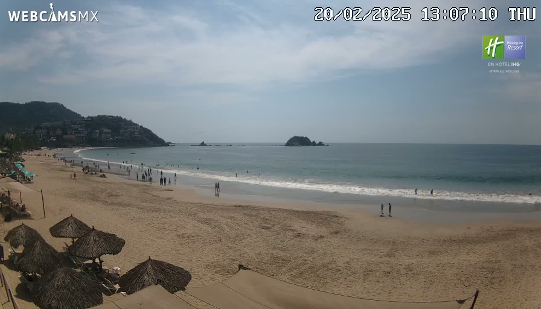 A sandy beach with calm ocean waves, under a mostly sunny sky, features scattered beachgoers and several thatched umbrellas near the shoreline, backed by hills with buildings visible in the distance.