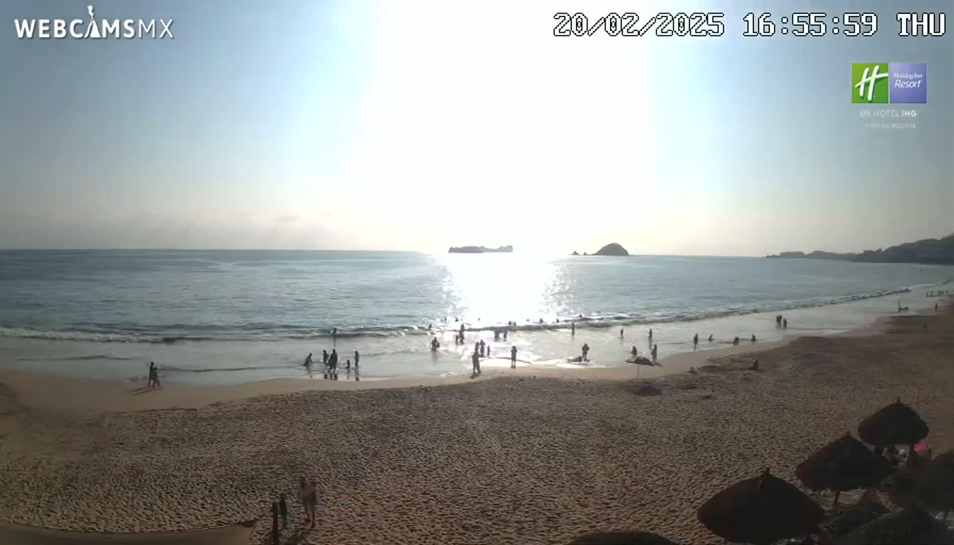 A sandy beach with people swimming and relaxing in the shallow water, under a bright, sunny sky, with islands visible in the distance.