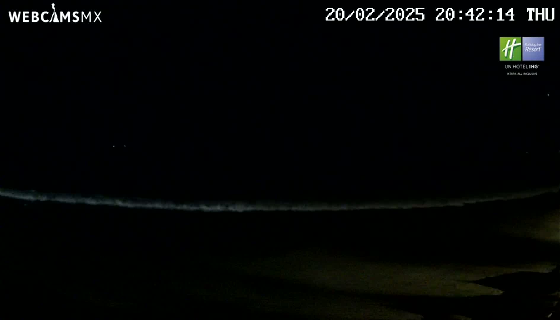 A dark nighttime beach scene shows a faint line of white waves breaking on the shore under a clear, black sky.