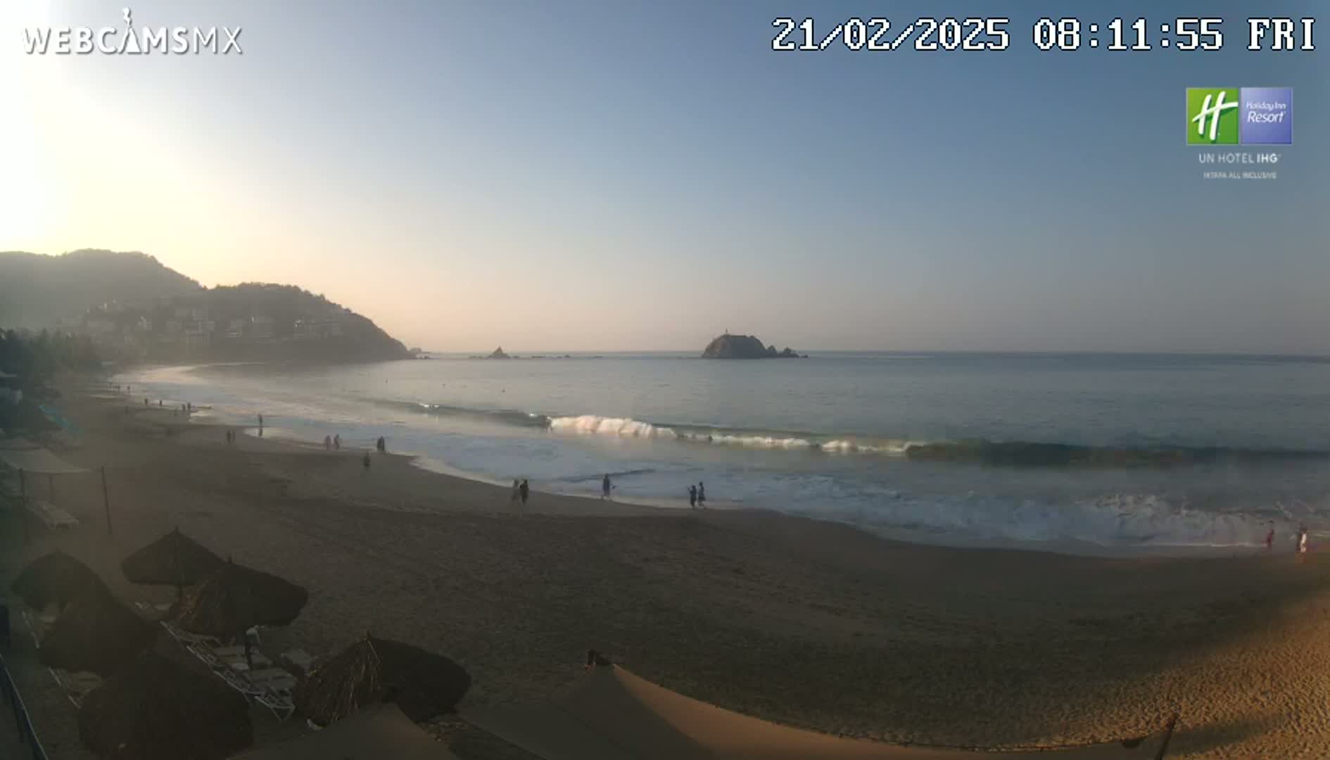 A sandy beach with gentle waves laps the shore under a clear, sunrise sky, with hills and a small island visible in the distance.