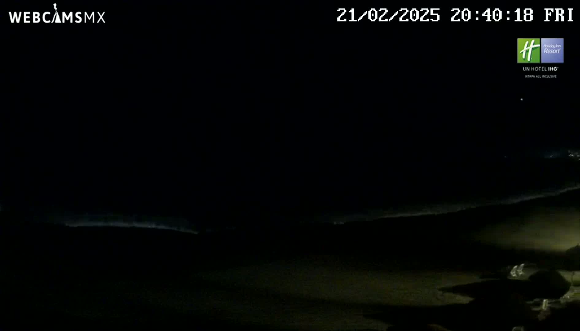 A dark nighttime beach scene shows a faint line of waves on the ocean under a clear, black sky.