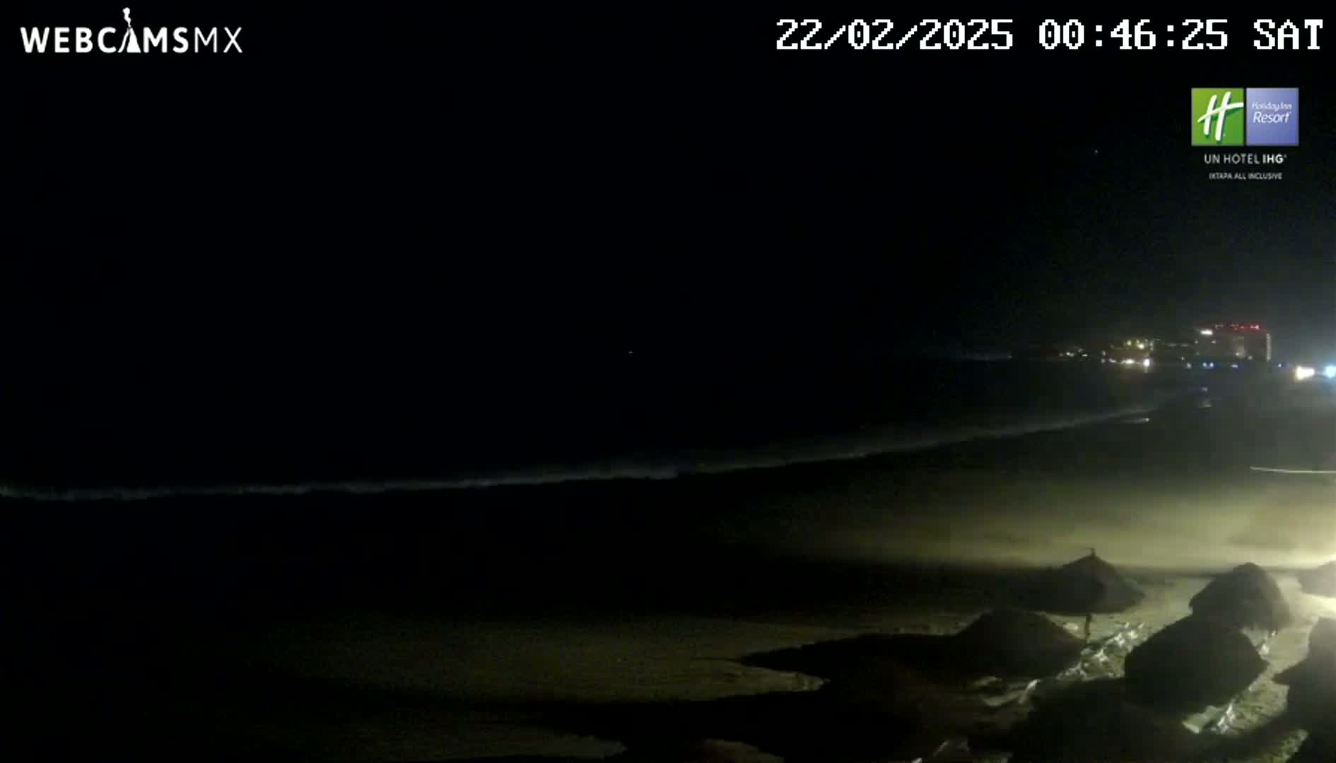 A dark, nighttime beach scene shows gentle waves lapping the shore under a clear, starless sky, with several dark beach umbrellas visible in the foreground.